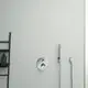 Einzelthermostat in Chrom von Ideal Standard an der Wand, mit einer Handbrause und einem an der Wand montierten Schlauch. In der Nähe ein schwarzes Regal mit Pflegeprodukten.